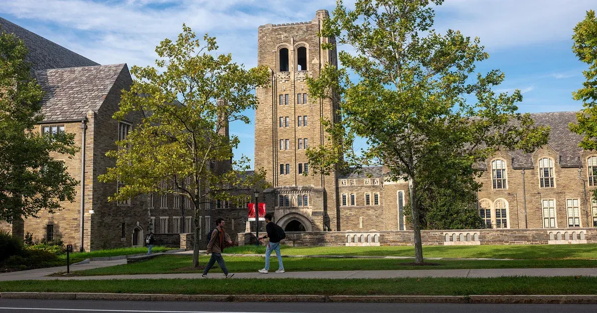 Stone Gothic campus buildings and tower with students walking on a sunny lawn path.