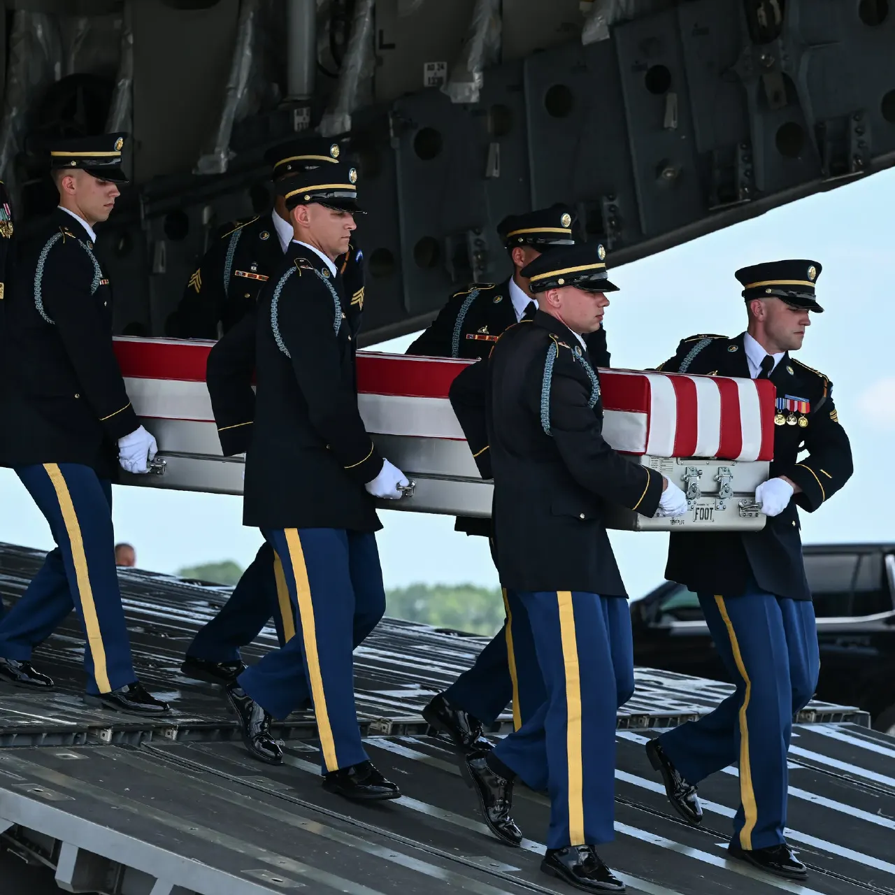 Uniformed honor guard carries a flag-draped casualty coffin off a military transport aircraft.