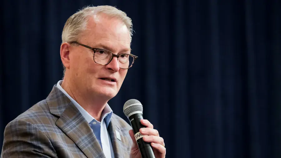 Man in glasses and plaid blazer speaking into a handheld microphone against a dark curtain.