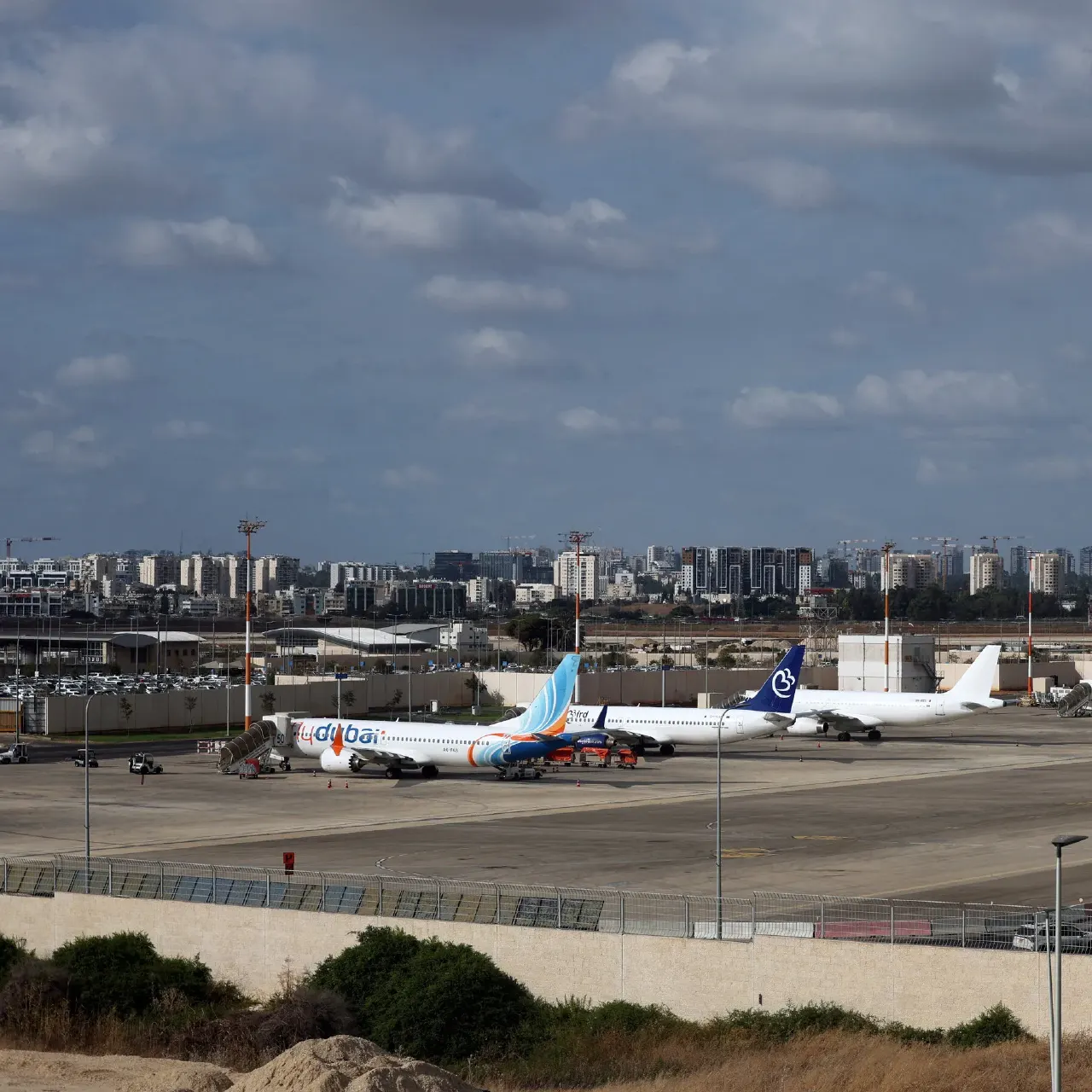 Flydubai jet parked on airport apron beside another airliner, city skyline behind.