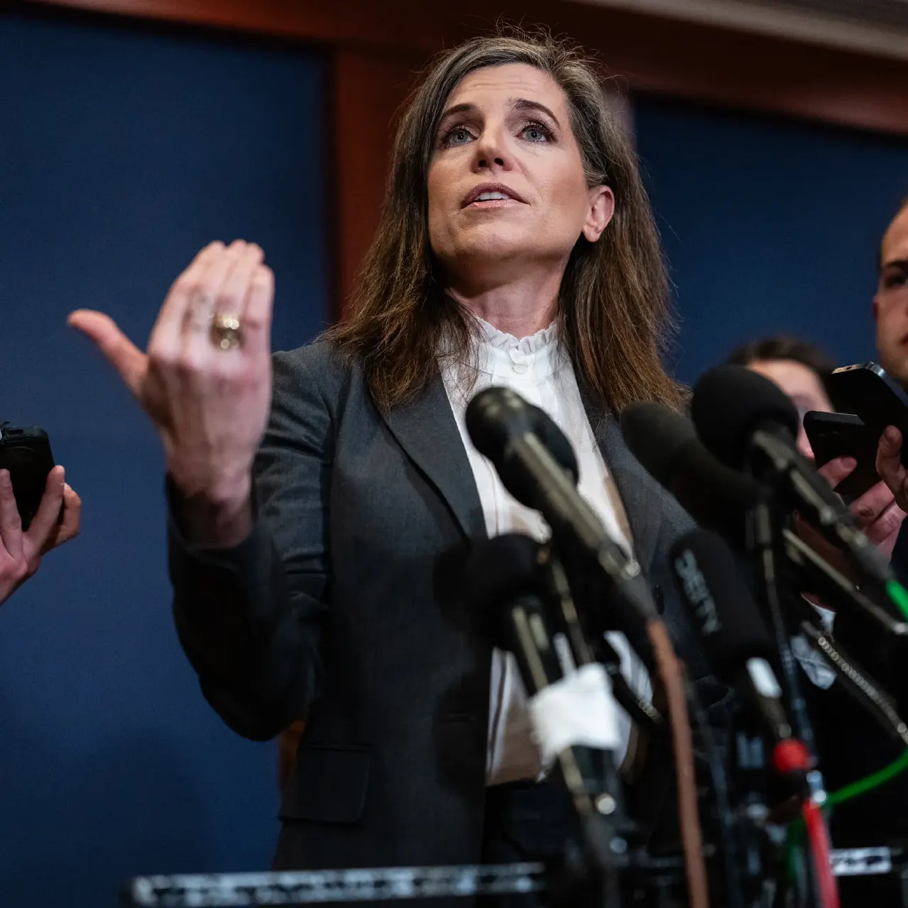 A woman in a dark blazer speaks at a press conference, gesturing before a bank of microphones.