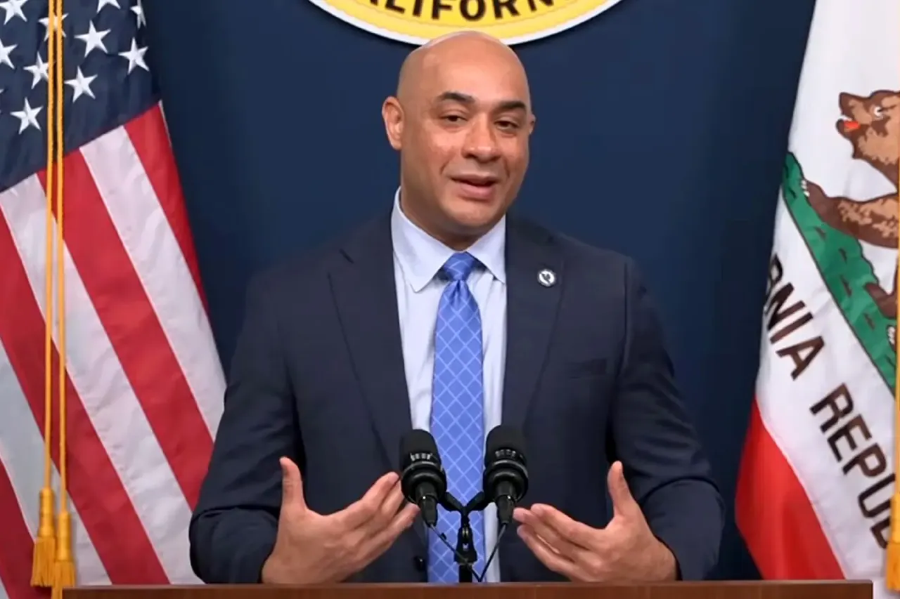 A bald man in a suit speaks at a podium before American and California flags.