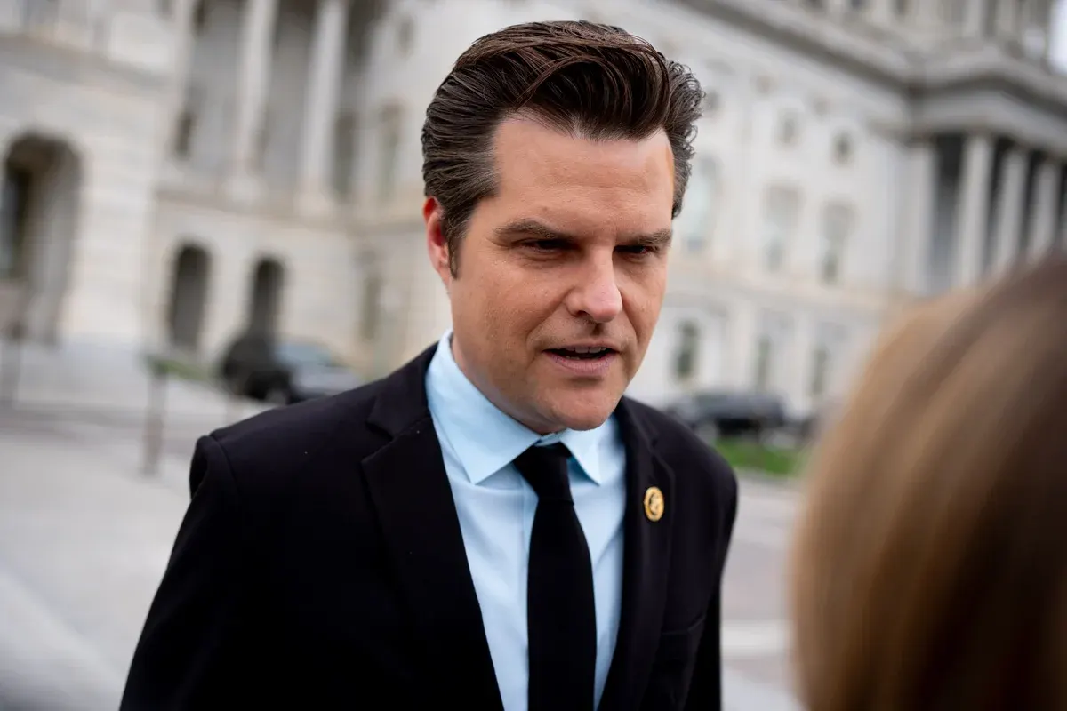 Man in dark suit and tie with lapel pin speaking outdoors near a columned government-style building.