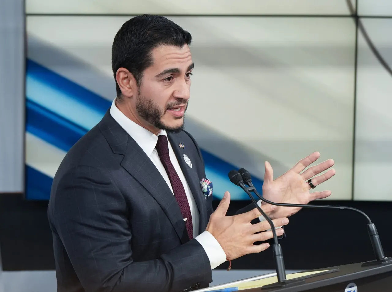 Man in dark suit gestures while speaking at a podium with microphones.