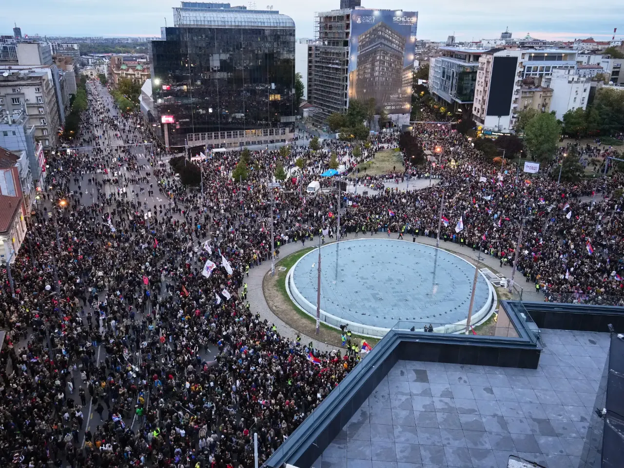 Aerial view of a huge crowd filling a Belgrade square around a circular fountain, with flags.