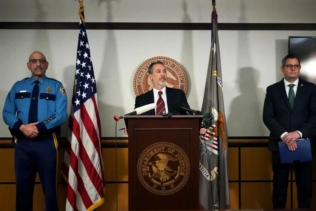 Official at podium before Department of Justice seal and flags, with uniformed and suited men beside him.
