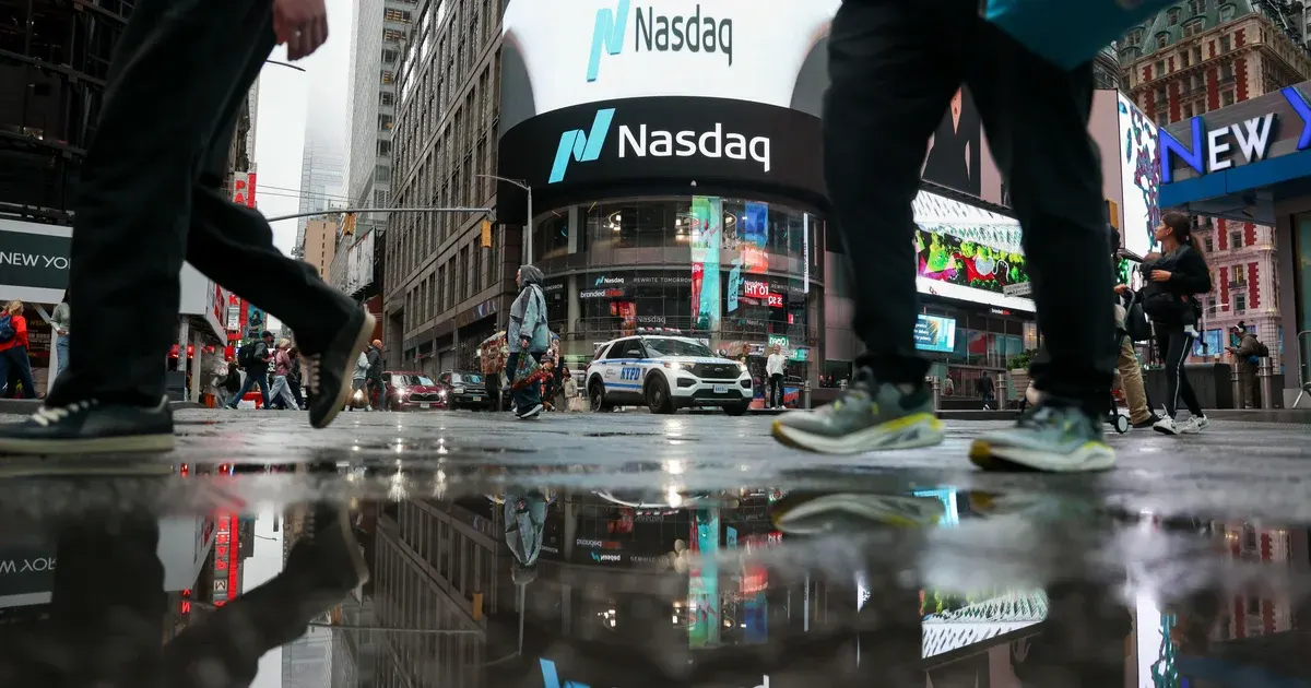 Pedestrians pass the Nasdaq MarketSite screens in Times Square on a wet day, reflected in puddles.
