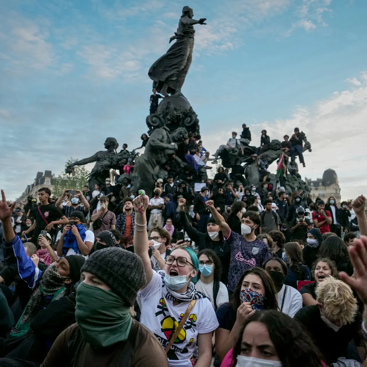 Large crowd of masked young people gathered around a bronze statue in an open plaza, some raising fists.