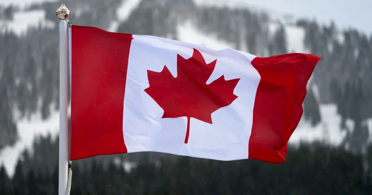 Canadian flag waving on a pole in front of snowy evergreen-covered mountains.