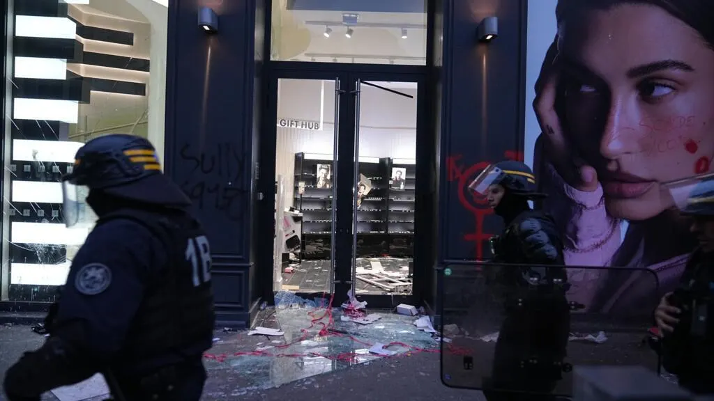 Riot police in helmets stand outside a shop with smashed glass and debris scattered on the pavement.