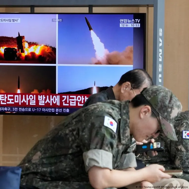 Soldiers in camouflage watch TV news showing missile launches with Korean ticker text.