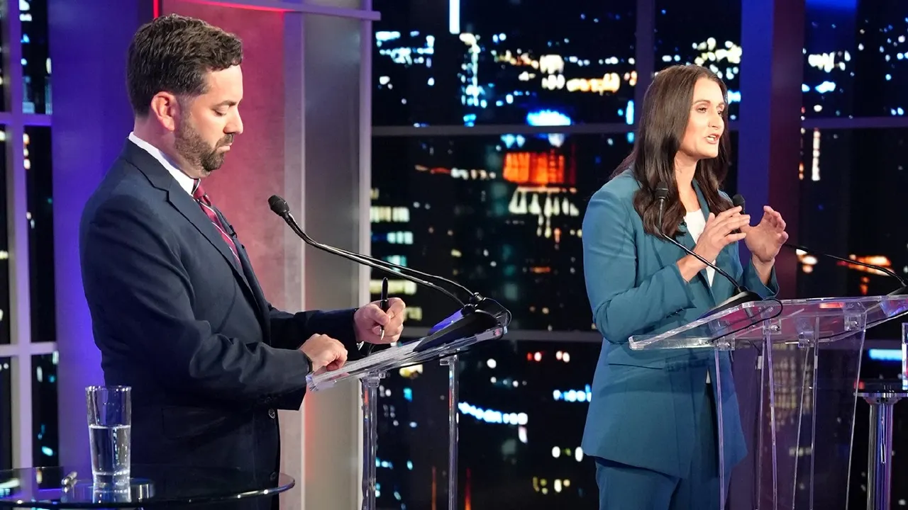 Two debate candidates stand at clear podiums on a studio set, one looking down, one gesturing.