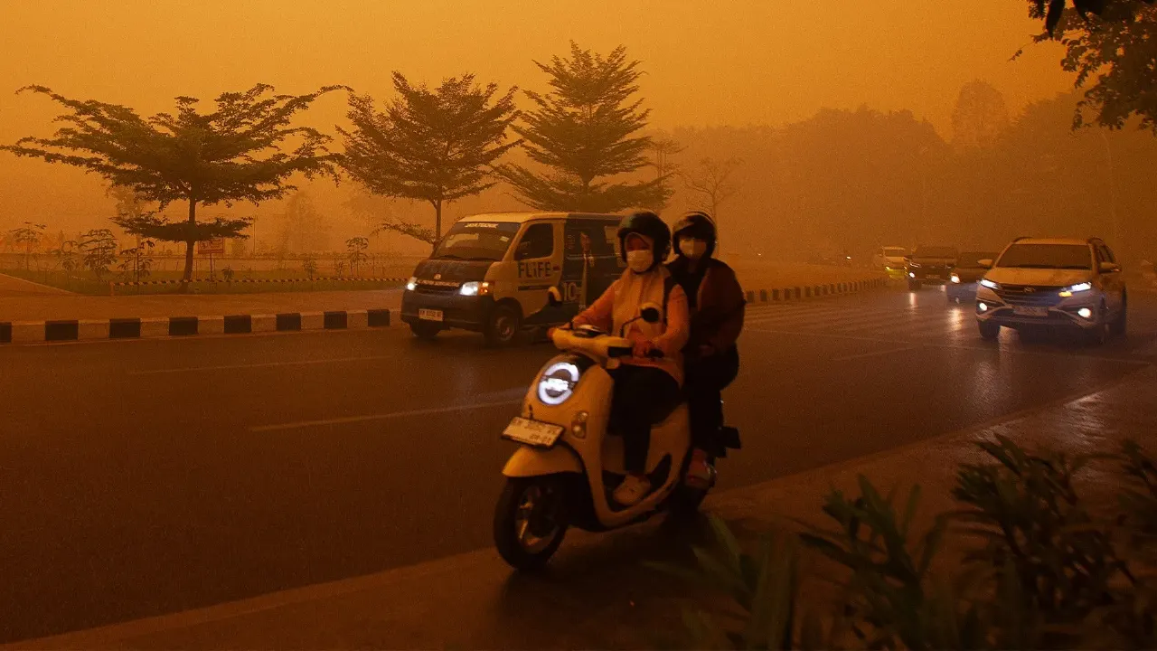 Two masked riders on a scooter on a street, with cars, under an orange smoke-filled sky.