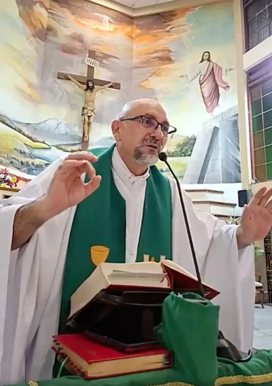 Priest in white vestment and green stole speaking at a microphone beside a painted church mural with crucifix.