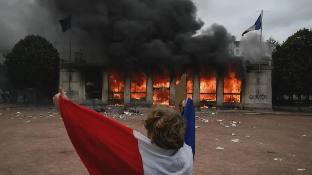 Protester with French tricolour flag watches a building burn behind a smoky square.