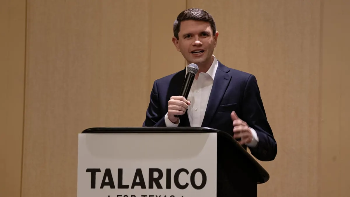 A man in a navy blazer speaks into a microphone at a podium bearing a campaign sign.