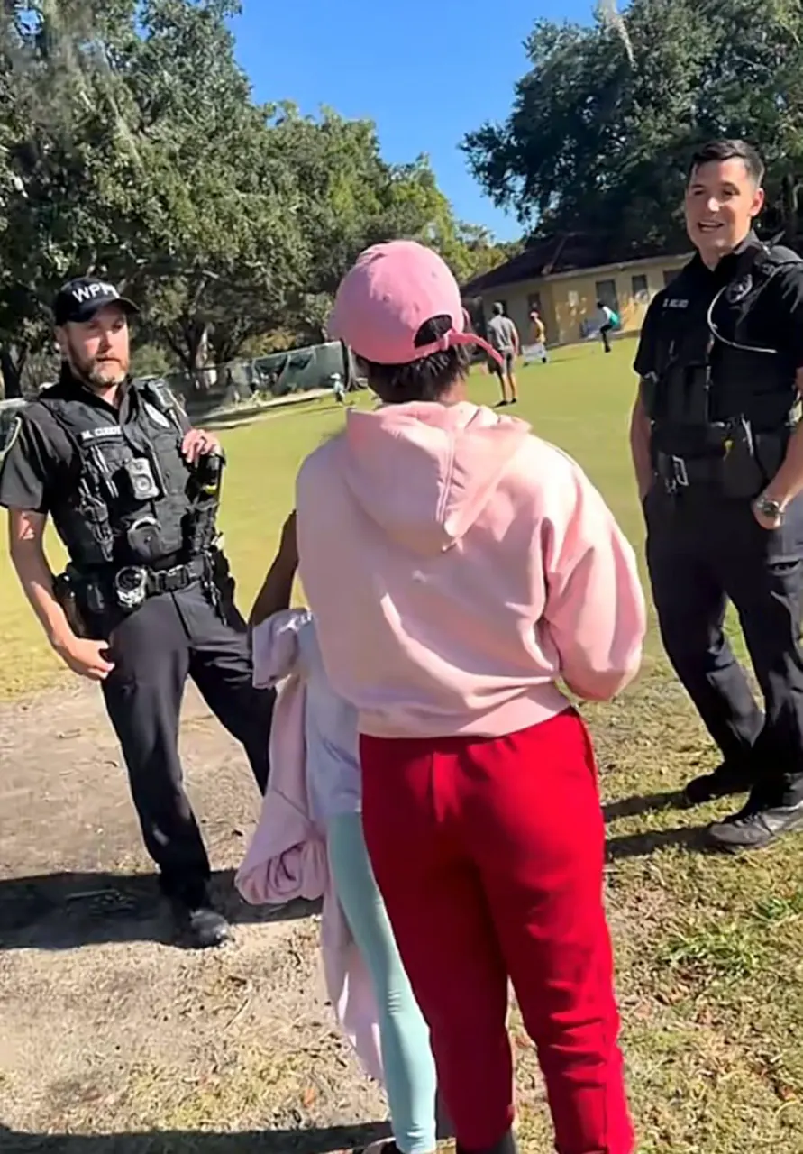 Two uniformed officers talk with a person in a pink cap and hoodie on a grassy field, with a young child visible.