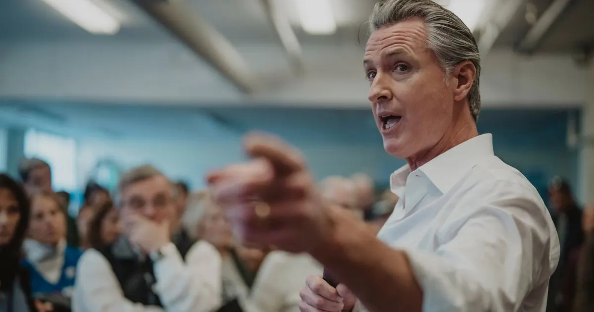 A gray-haired man in a white shirt gestures while speaking to a seated audience indoors.