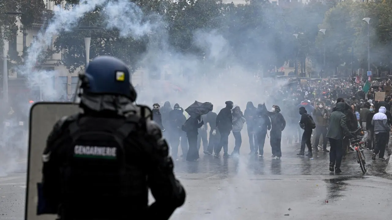 Riot police in helmets and gear watch a crowd through tear gas smoke on a city street.