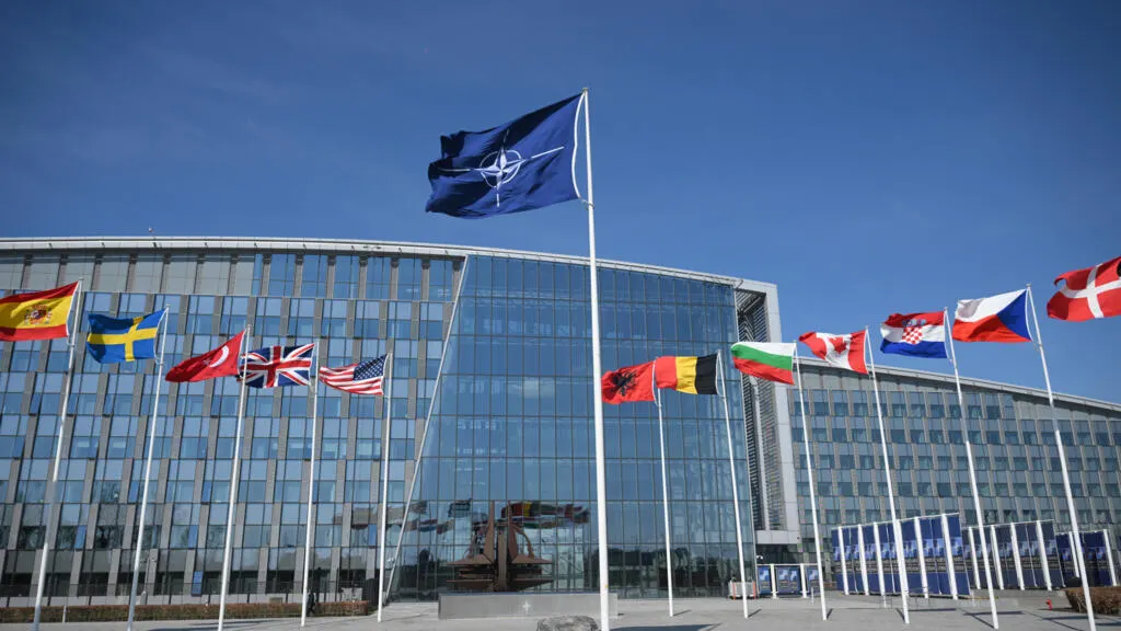 NATO flag and member nations' flags flying before a large glass government-style building