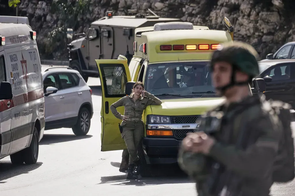 Uniformed soldier with a helmet and rifle in foreground; woman soldier beside a yellow ambulance on a road.