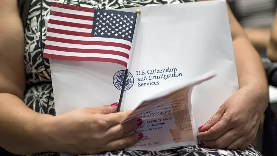 Person holding U.S. Citizenship and Immigration Services envelope with small American flag and naturalization documents.
