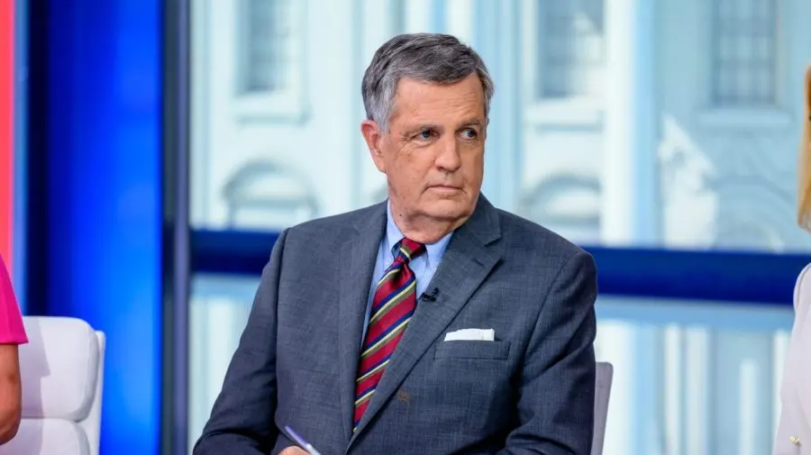 Older man in grey suit and striped tie seated on a studio set, looking off-camera.
