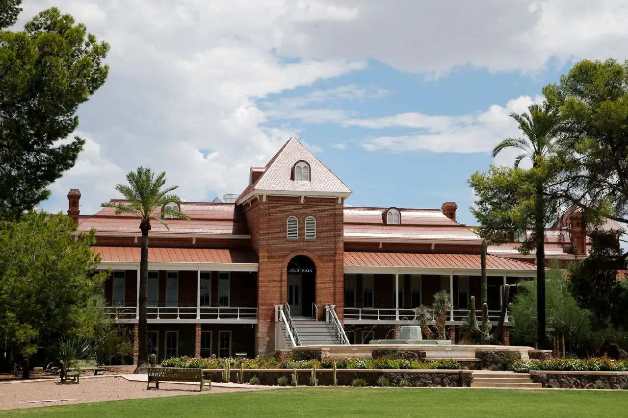 Historic red brick university building with palm trees, lawn, and benches under a cloudy sky.