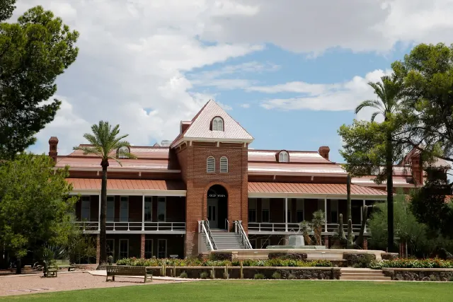 Historic red brick university building with palm trees, lawn, and benches under a cloudy sky.
