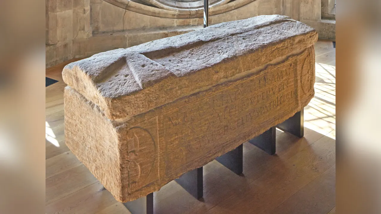 Carved stone Roman sarcophagus with inscription displayed indoors on wooden floor.