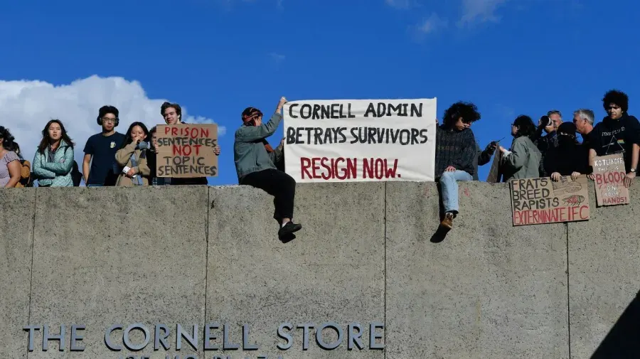 Students on a concrete wall protesting with banners reading 'Cornell Admin Betrays Survivors, Resign Now'