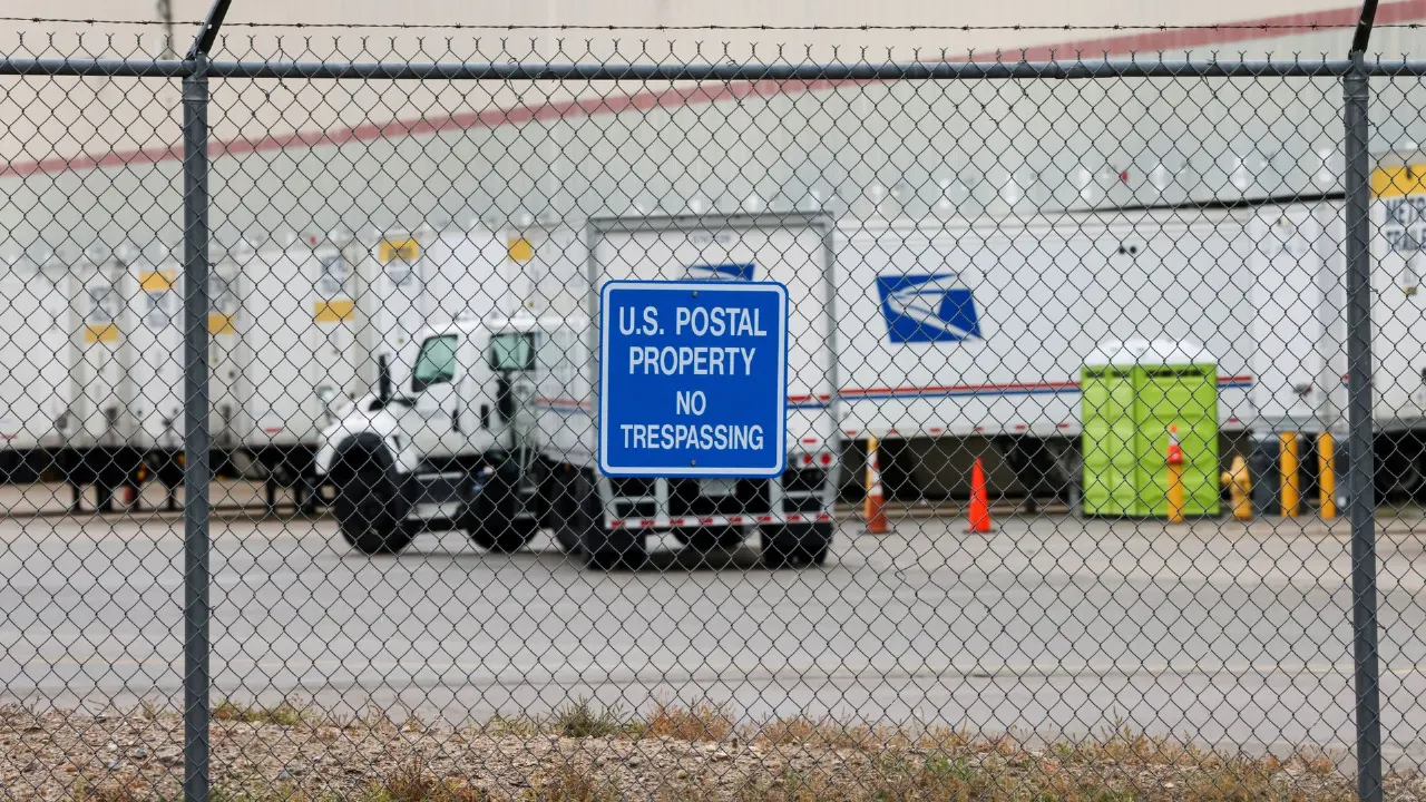 Chain-link fence before US Postal property sign, postal trucks and USPS eagle logo behind.