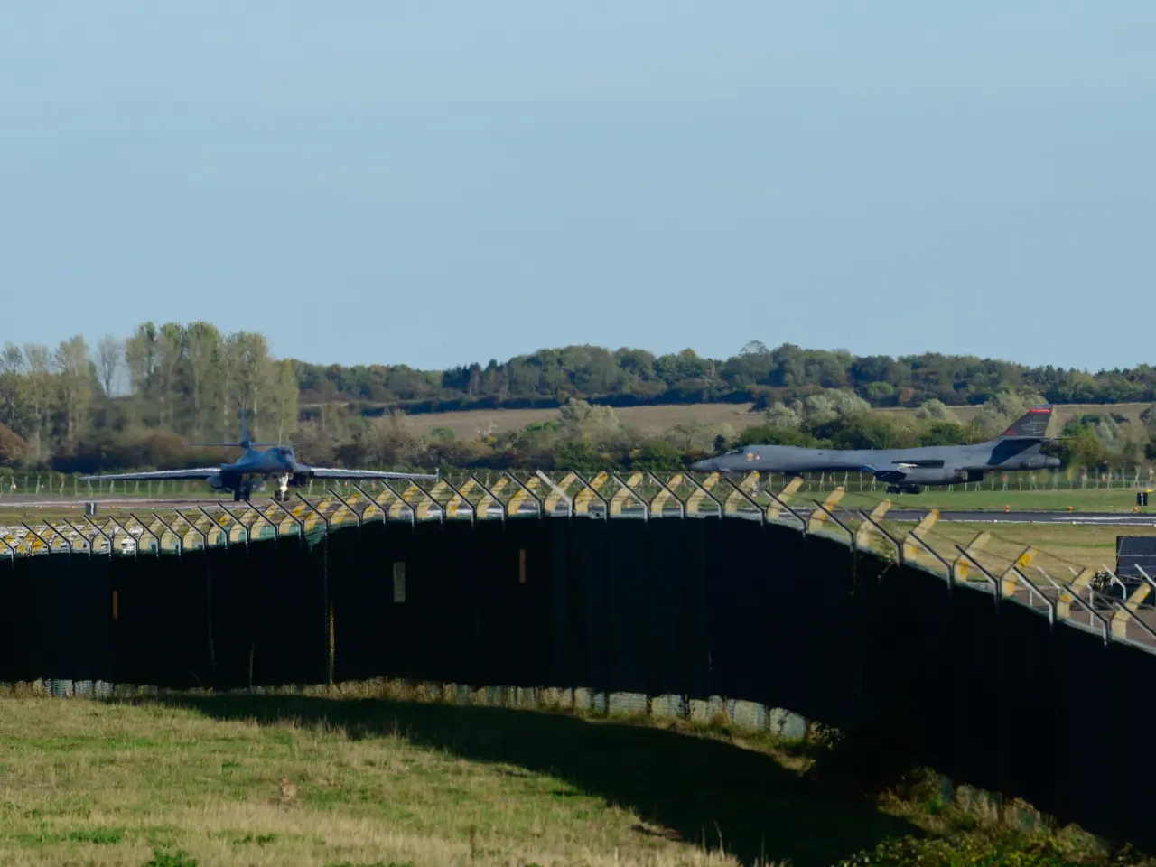 Two military bombers on an airfield runway behind a long perimeter fence under a clear sky.