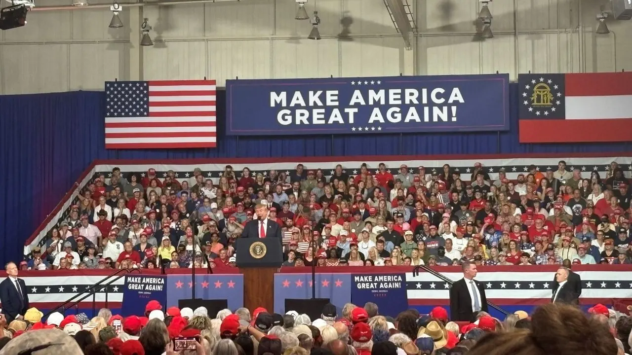 Speaker at a podium before a large crowd in red caps under Make America Great Again banners and flags.