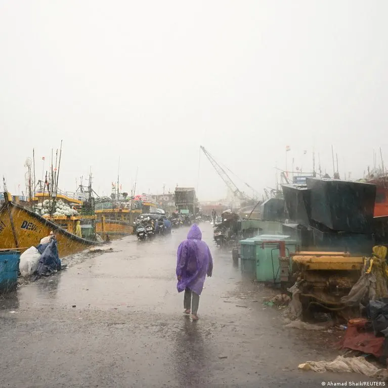 Person in purple raincoat walks through heavy rain past moored fishing boats at a harbour