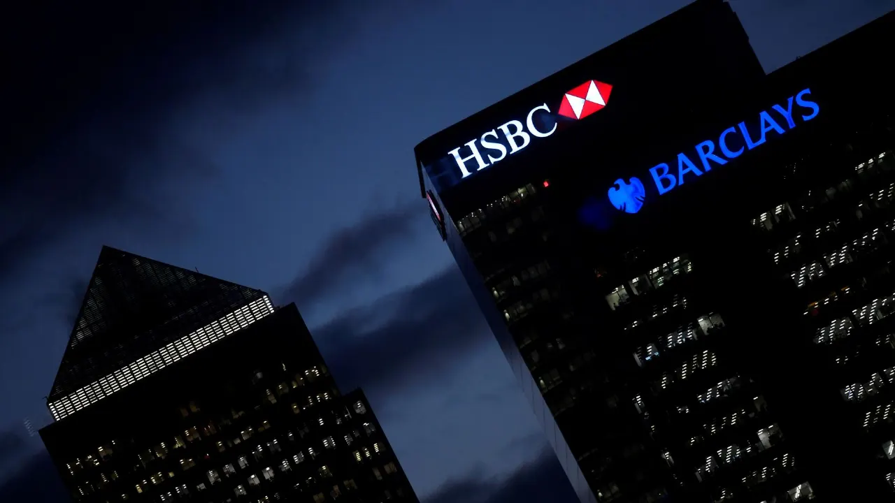 Dusk view of office towers displaying HSBC and Barclays bank logos at night.