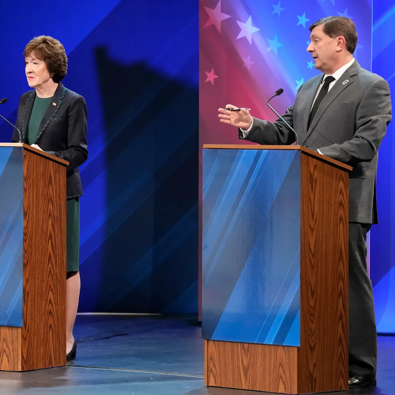 Two debate candidates stand at separate podiums on a blue stage, one gesturing while speaking.