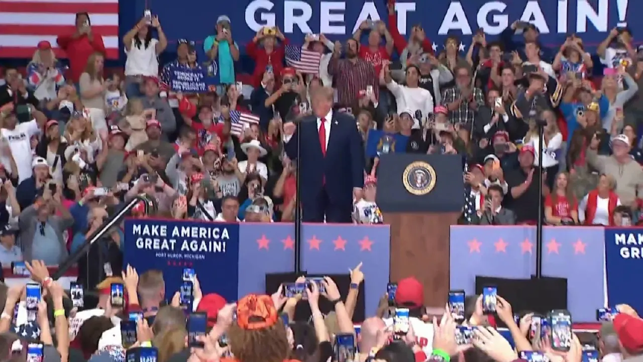 Large rally crowd filming a speaker at a podium under 'Make America Great Again' banners, with American flags.