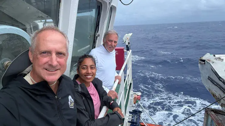 Three people smile on a ship deck at sea, with waves and a lifeboat visible.