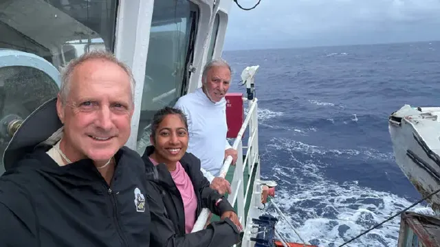 Three people smile on a ship deck at sea, with waves and a lifeboat visible.