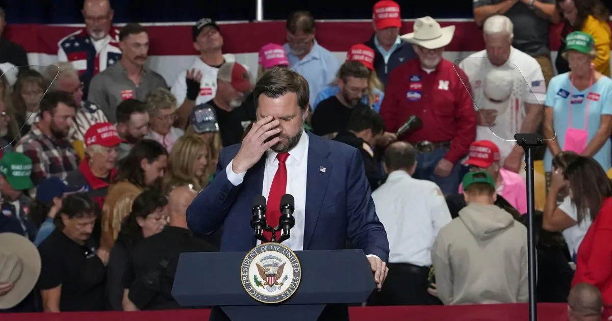 A man in a navy suit pauses at a podium bearing a presidential seal, hand to his head, before a crowd.