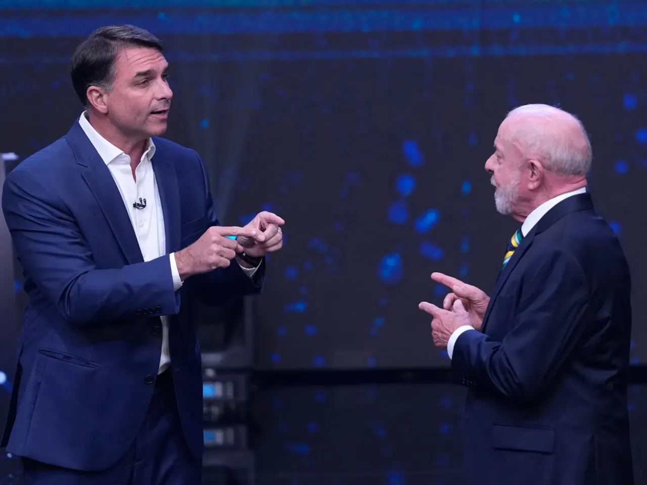 Two men in suits gesture at each other during a televised debate on a dark stage.
