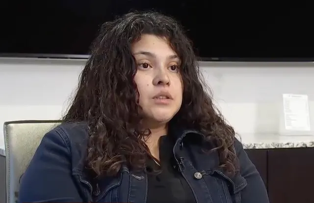 Woman with long curly dark hair in denim jacket speaking during a television interview.