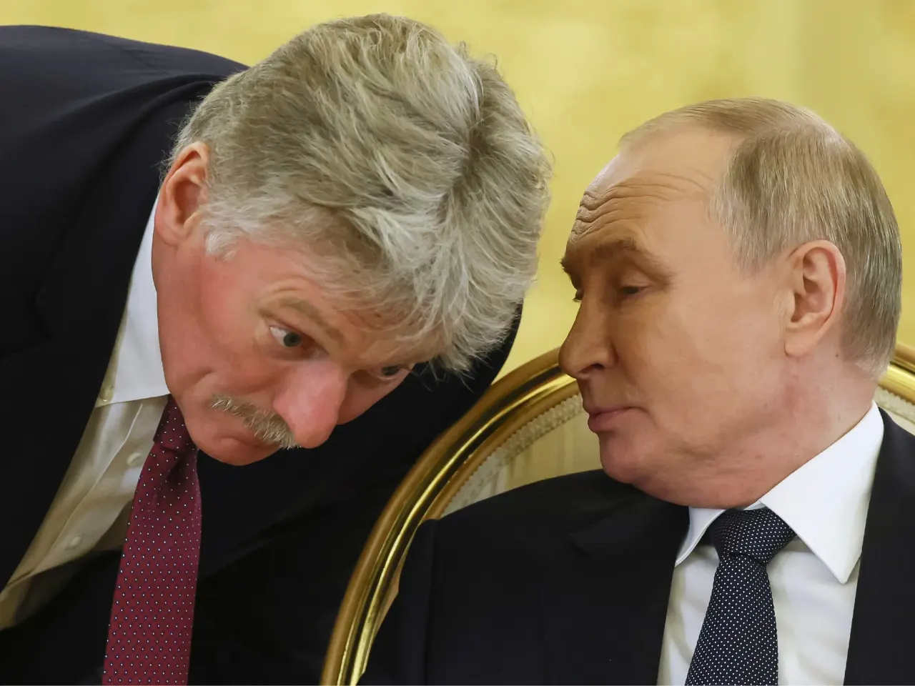 Two men in dark suits in close conversation, one leaning in, gilded chair background.