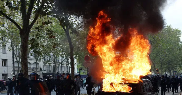 Riot police in helmets stand near a large fire and burning car on a Paris street.