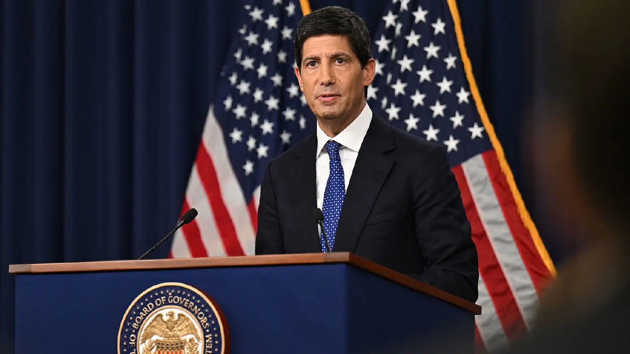 Man in dark suit speaking at podium with Federal Reserve seal, flags behind.