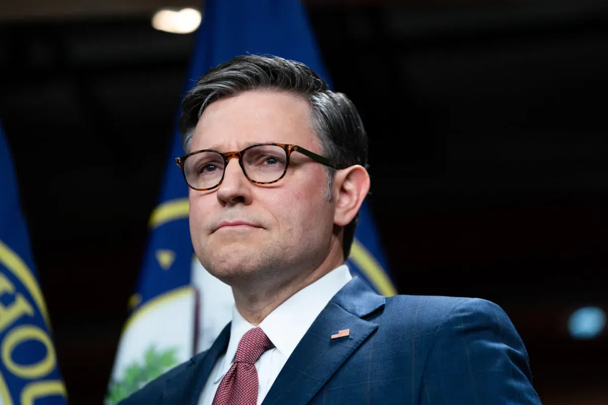 Man in glasses and dark suit with pin, blue flags behind, looking off-camera.