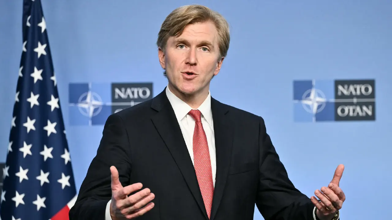 Man in dark suit and red tie speaking at press event before NATO and US flags.