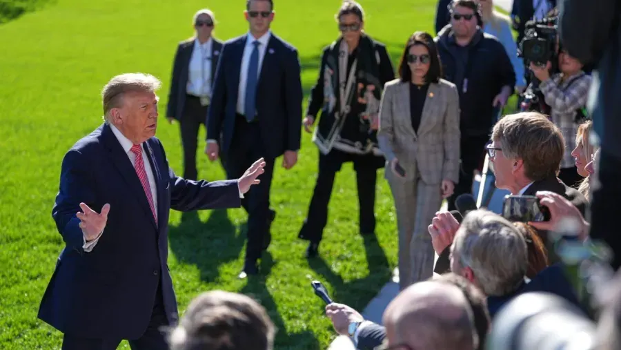 A man in a navy suit gestures while speaking to reporters on a sunny lawn, with staff and press nearby.