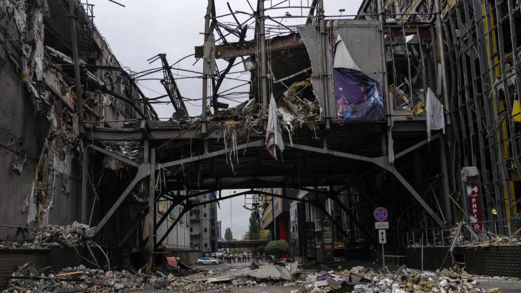 Bombed-out shopping centre with collapsed steel framework and rubble in a city street.